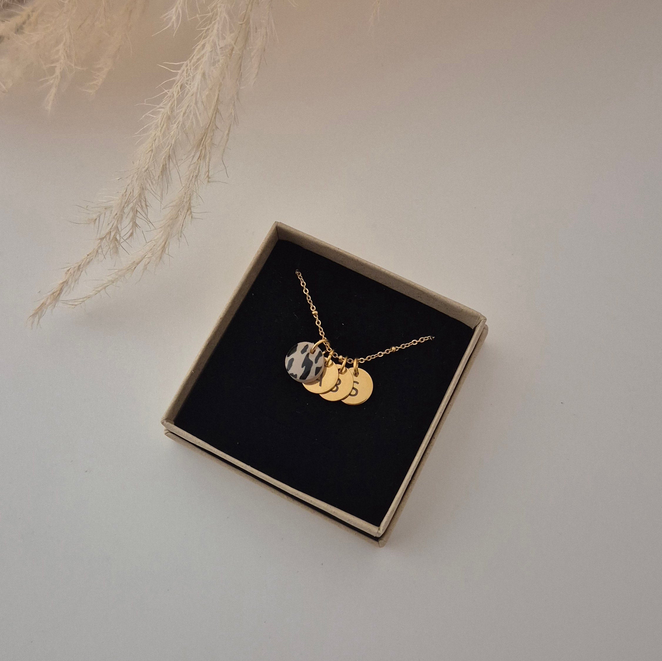 Die goldene Kette ist in der Schmuckschachtel mit dem Leo-Anhänger und 3 goldenen Plättchen-Anhängern, mit jeweils unterschiedlichen Buchstaben, welche nicht so gut erkennbar sind, da sie leicht übereinander liegen.