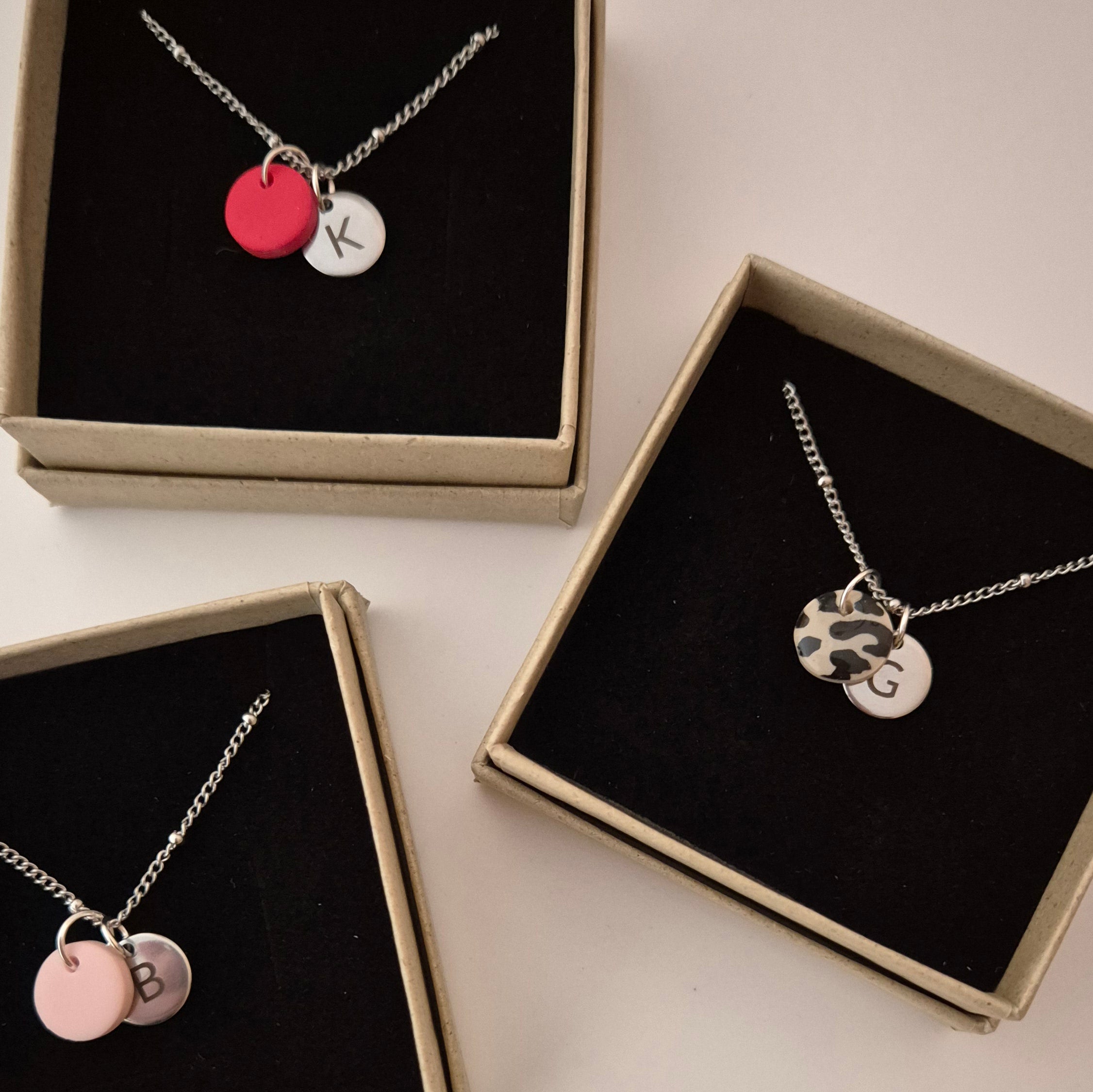 Nähere Aufnahme der Ketten. Drei Ketten in silber mit der gleichen Punkt-Struktur wie die goldene Kette. Die Ketten liegen in jeweils einer Schmuckschachtel und haben jeweils einen anderen Anhänger in rot, rosa und Leo. Jede Kette hat außerdem einen silbernen Anhänger mit Gravur in den gleichen Größen wie die goldene Kette.