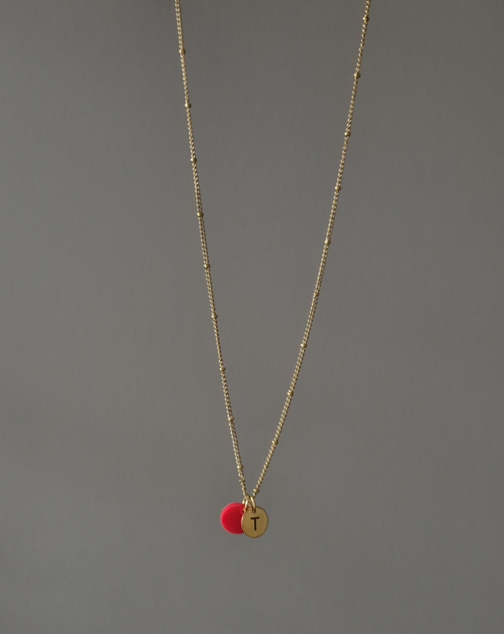 Nahaufnahme der hängenden Kette in gold. Die Kette ist fein und hat ca. alle 2 cm einen kleinen goldenen Punkt, was die Kette aufregender gestaltet. Die Kette ist mit einem roten Plättchen (ca. 1cm) versehen sowie einem gravierten Plättchen in rund und gold mit einem gravierten, schlichtem T.