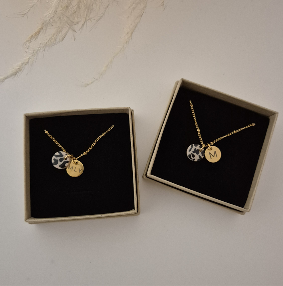 Zwei goldene Ketten in einer Schmuckbox, beide Ketten haben den beige/schwarzen Leo Anhänger (1cm) sowie den goldeen Anhänger mit Gravur (1cm). Der linke Anhänger hat eine Gravur mit 3 Buchstaben &