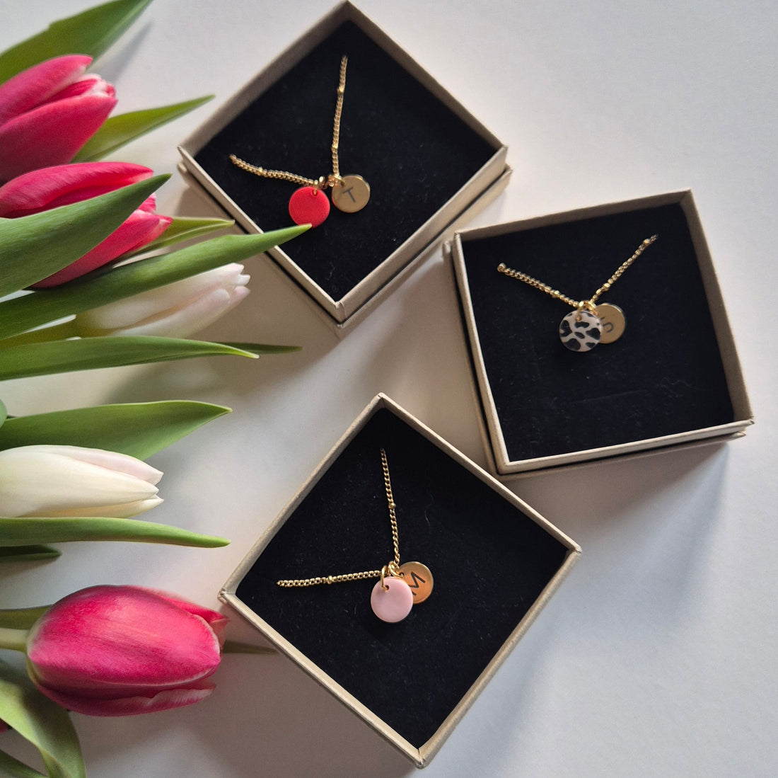 Es liegen die drei Ketten mit den unterschiedlichen Anhängern in rot, Leo und rosa plus das gravierte Plättcen in jeweils einer Schmuckbox. Zur Dekoration befinden sich daneben Tulpen.