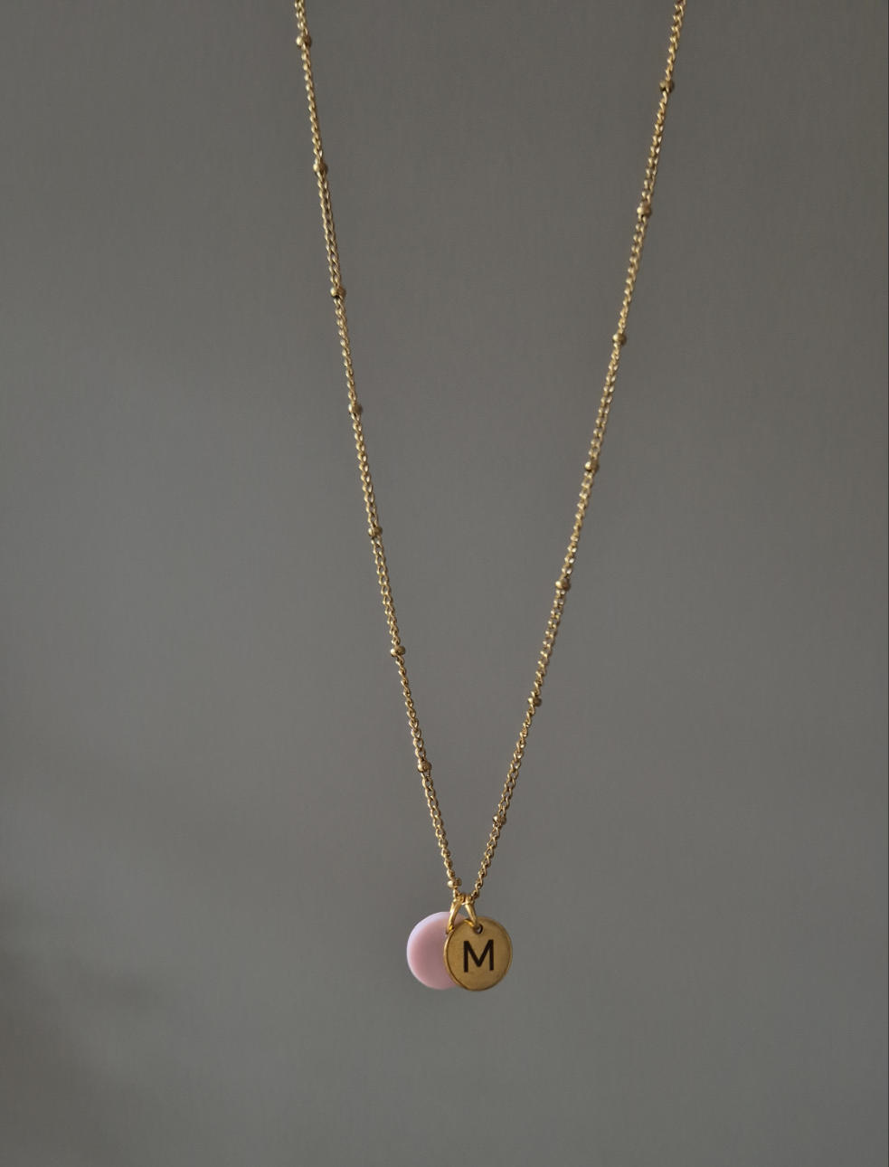 Nahaufnahme der hängenden Kette in gold. Die Kette ist fein und hat ca. alle 2 cm einen kleinen goldenen Punkt, was die Kette aufregender gestaltet. Die Kette ist mit einem rosanen Plättchen (ca. 1cm) versehen sowie einem gravierten Plättchen in rund und gold mit einem gravierten, schlichtem M.