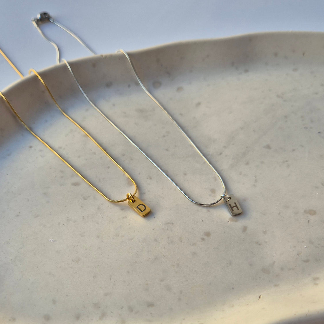 Liegende Ketten in silber und gold auf einer Schale. In der Nahaufnahme sieht man die rechteckigen Anhänger mit der Gravur.