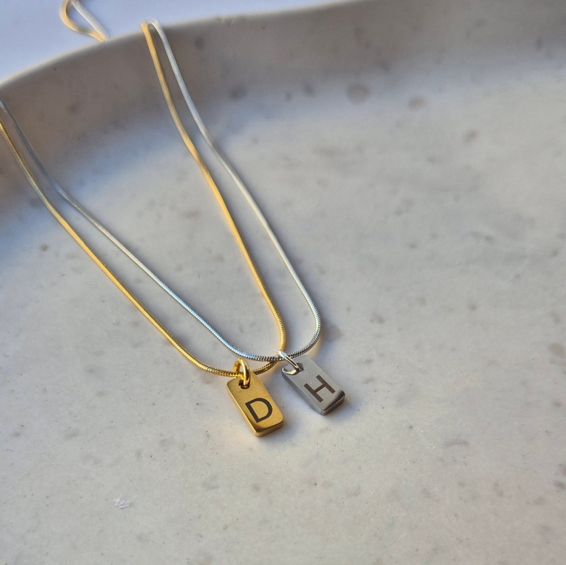 Feine goldene und silberne Kette mit je einem kleinen, rechteckigen Anhänger in der gleichen Farbe. Auf dem Anhänger ist jeweils ein einzelner Buchstabe graviert. Die Kette hat eine glatte, filigrane Struktur und einen sanften Glanz.
